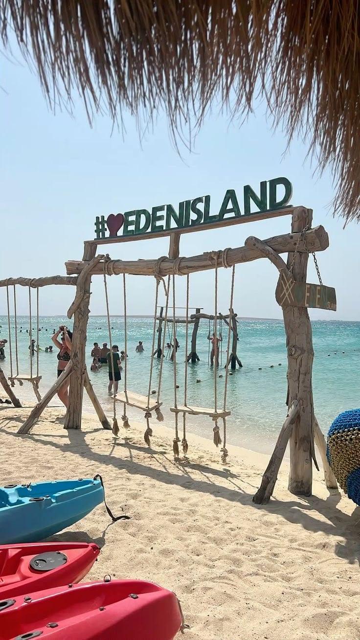 Wooden swings under the Eden Island sign by the turquoise water, with kayaks on the sand