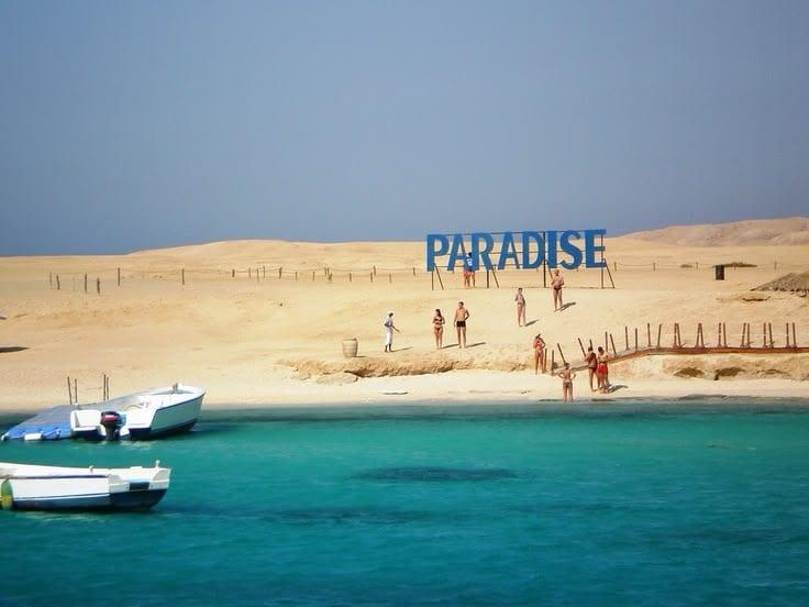 Paradise sign on the sandy island shore with boats in the turquoise water